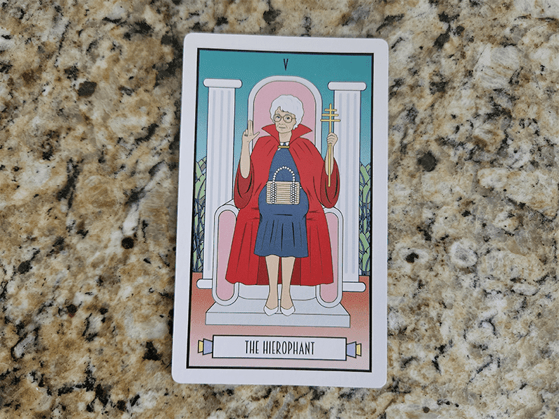 Sophia Petrillo wearing a blue dress and red cape, sitting on a throne framed by two columns, and holding up a cross on The Hierophant card.