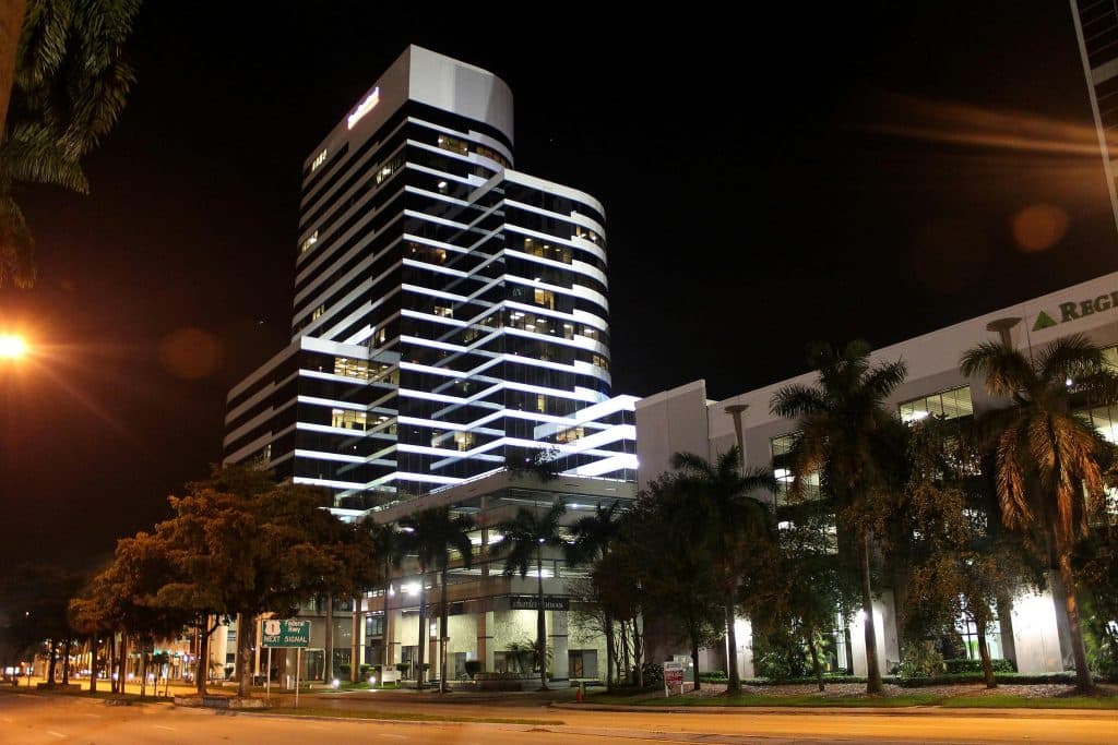 A modern skyscraper illuminated at night with palm trees lining the street in an urban setting.
