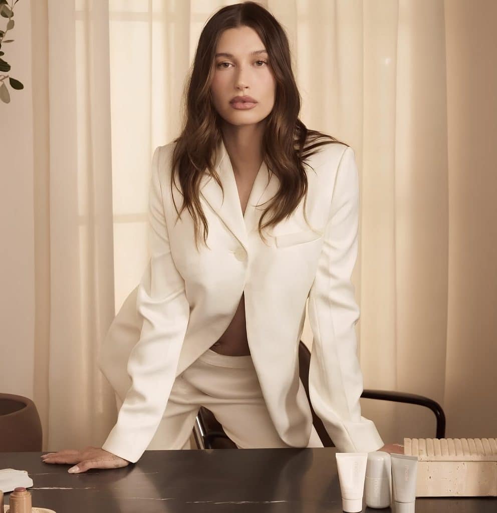 Hailey Bieber in an offwhite pant suit leaning forward on a brown table with Rhode beauty products on it.