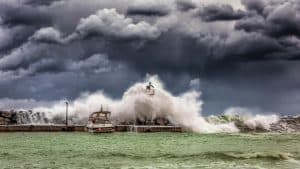 Powerful waves of one of worst hurricanes crash against a pier and lighthouse under dark stormy skies.