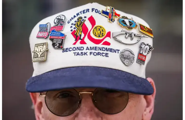A man wearing a white cap with a blue brim, adorned with various NRA and Second Amendment pins. The cap reads "Charter Founding Member, Second Amendment Task Force." The individual wears sunglasses, conveying a serious tone. The NRA calls for a full investigation into the shooting of Alex Pretti.