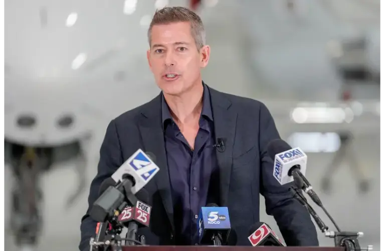 A man in a dark suit speaks at a podium with multiple microphones from news outlets. The background is blurred, suggesting an indoor setting. The FAA is still investigation the plane crash at Bangor International Airport.
