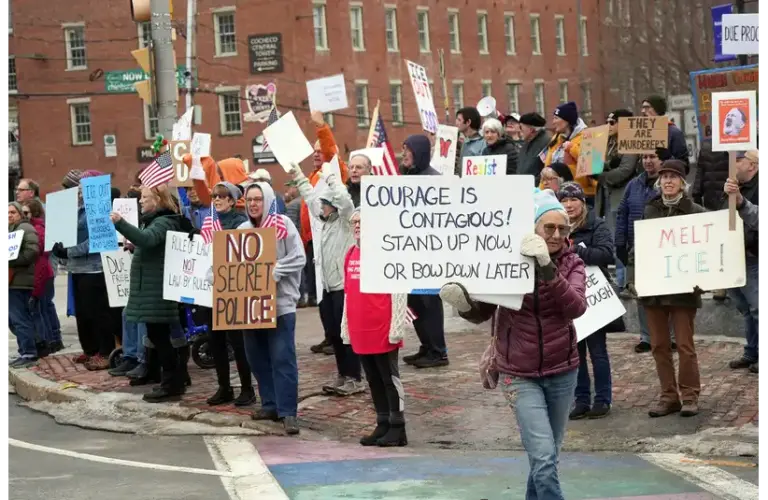 A diverse group of protesters in Minneapolis stand on a street corner, holding signs with messages like "Courage is Contagious" and "No Secret Police." The atmosphere is determined and spirited at the Minneapolis protests.