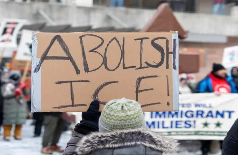 Person in winter clothing holds a cardboard sign reading "Abolish ICE!" at a protest. Background shows blurred crowd and banners in a snowy setting.