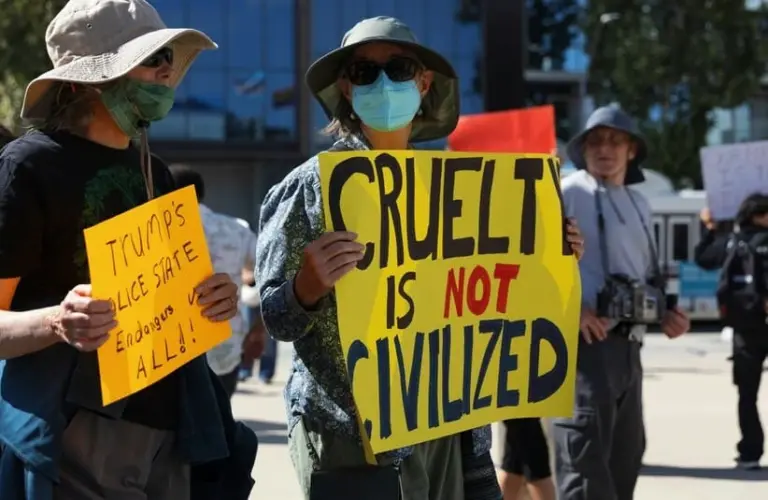 Protesters against ICE wearing hats and masks hold signs reading "Cruelty is Not Civilized" and "Trump’s Police State Endangers All" at an outdoor rally.