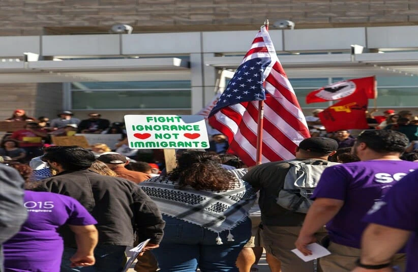 A diverse crowd gathered at a protest against ICE, holding a large American flag and a sign saying "Fight ignorance not immigrants" with red hearts. The mood is determined.