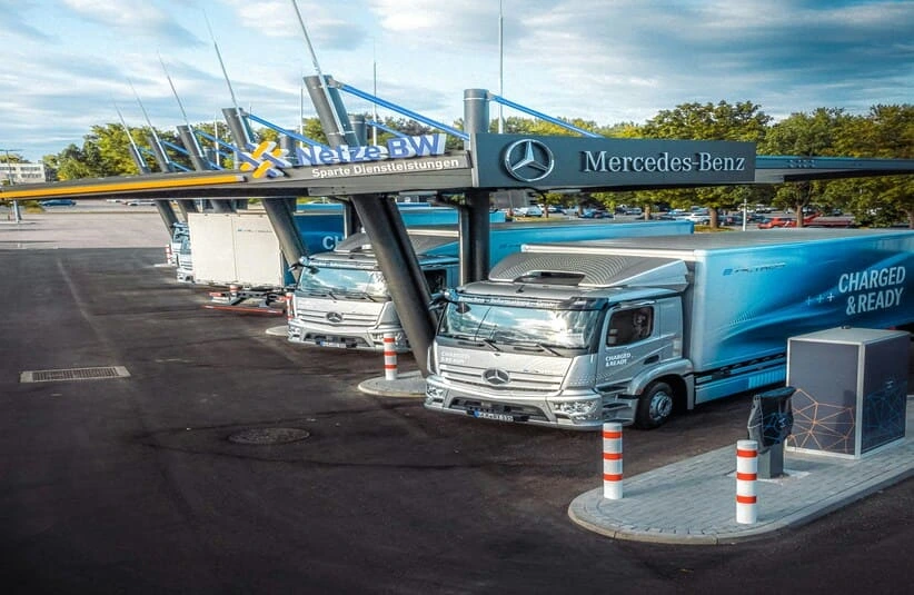 Three Mercedes-Benz electric trucks parked at a charging station under a cloudy sky. The trucks are labeled "Charged & Ready," with trees in the background.