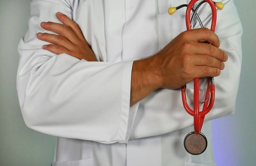 A doctor in a white coat crosses their arms, holding a red stethoscope. The setting conveys professionalism and healthcare. The tone is calm and confident.
