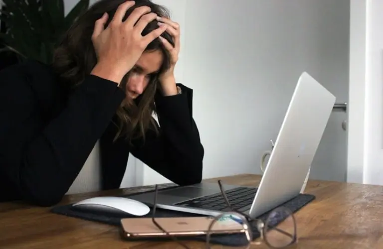A person with burnout sits at a wooden desk, head in hands, looking stressed. A laptop, mouse, phone, and glasses are on the table, conveying frustration.