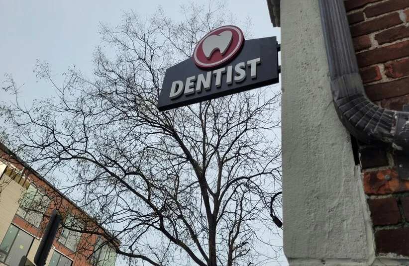 Ohio Dentist sign with a tooth icon on a building exterior. A bare tree and part of a brick building are visible against a cloudy sky. Urban setting.
