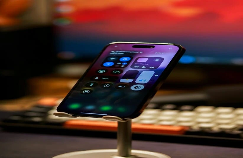 Close-up of an Apple iPhone on a stand, displaying a control center interface. A blurred keyboard and a monitor with a warm gradient are in the background.