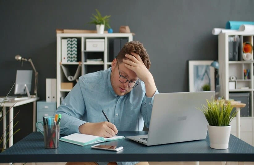 A worker experiencing burnout at their work desk. A man writing in a notebook in front of a laptop.