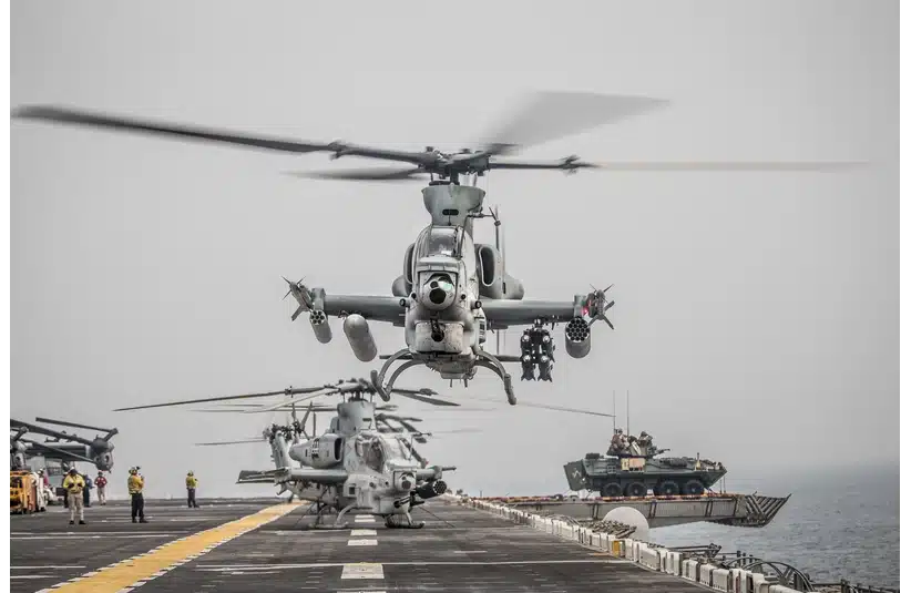 Helicopters land on an aircraft carrier in a foggy setting, with military personnel nearby and an armed vehicle on deck, conveying a tense atmosphere.