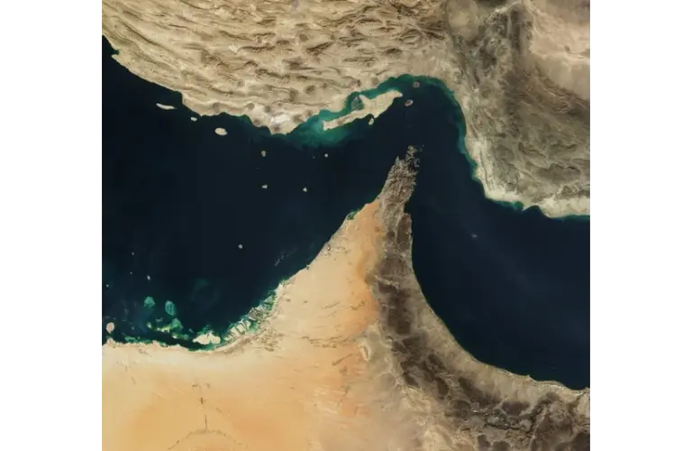 Satellite image of the Strait of Hormuz, featuring a dark blue body of water bordered by arid, sandy landscapes with rugged terrain.