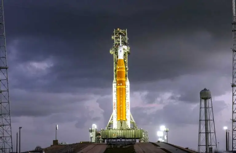 Artemis II rocket with an orange and white body stands on a launch pad against a dramatic, cloudy evening sky, illuminated by bright floodlights.