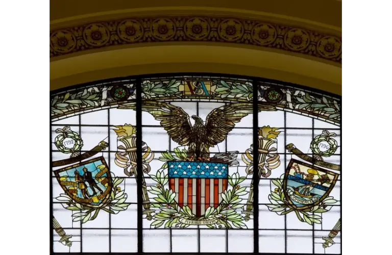 Stained glass window featuring an eagle and a shield, showcasing intricate colors and designs. Soon Congress will vote on the War Powers Resolution here.
