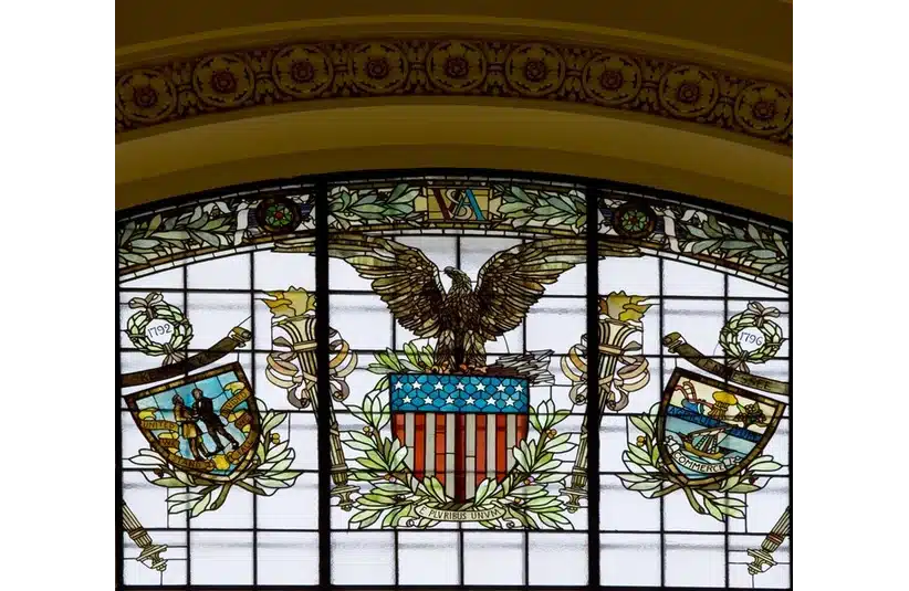Stained glass window featuring an eagle and a shield, showcasing intricate colors and designs. Soon Congress will vote on the War Powers Resolution here.