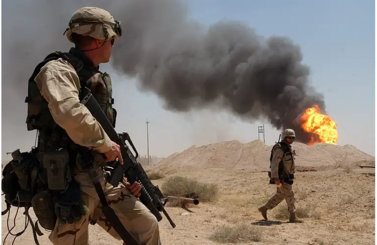 Two soldiers in desert camouflage gear stand on sandy terrain. One holds a rifle, and the other walks. Black smoke billows from a fire in the background. Showing similarities of the current U.S.-Iran crisis.