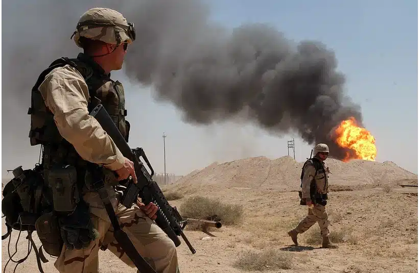 Two soldiers in desert camouflage gear stand on sandy terrain. One holds a rifle, and the other walks. Black smoke billows from a fire in the background. Showing similarities of the current U.S.-Iran crisis.