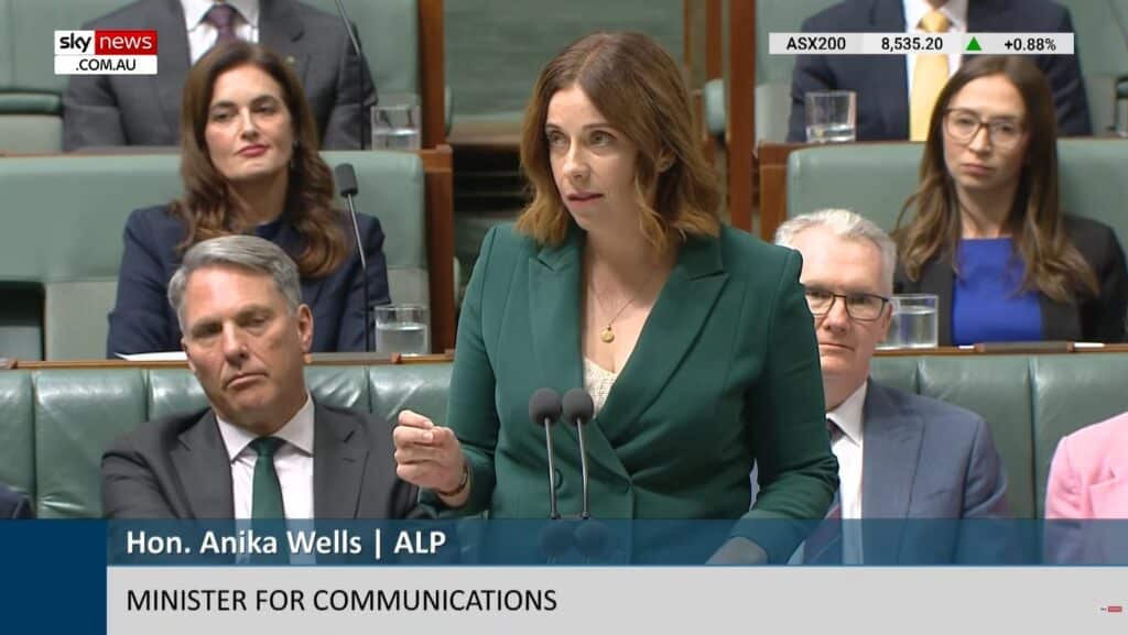A woman in a green suit speaks at a podium in Australian Parliament. People in professional attire are seated behind her, attentive. The tone is formal.