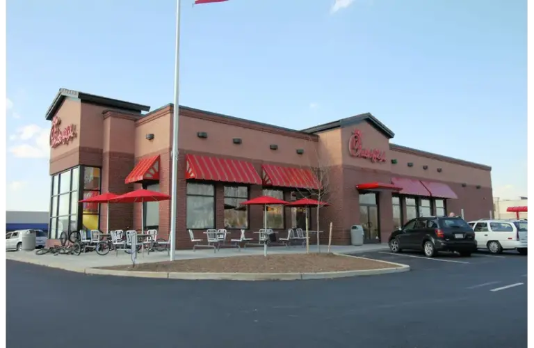 A Chick-fil-A restaurant with a red brick exterior and red striped awnings. The outdoor seating area has red umbrellas, and a few cars are parked nearby. NJ had a shooting at theirs.