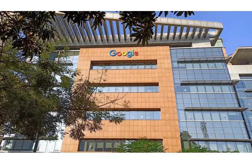 A multi-story Google office building with a mix of glass and brick facade, featuring the Google logo. Trees partially obscure the view, under a clear blue sky.