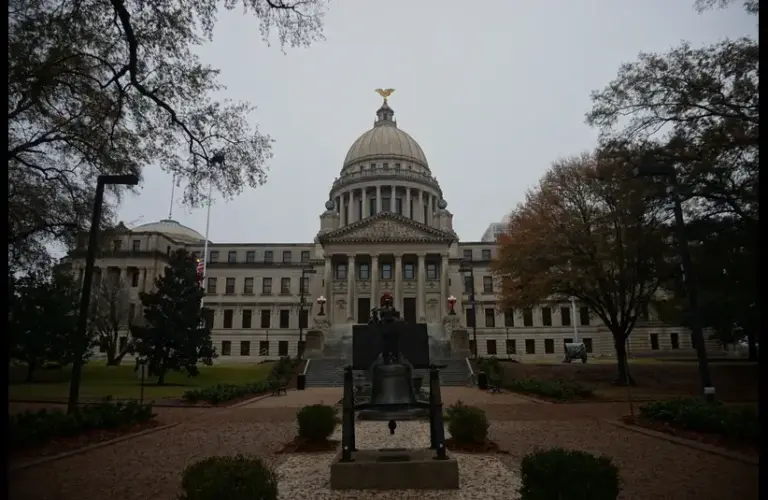 Mississippi State Capitol Building, a majestic government building with a large dome, surrounded by trees and a cloudy sky. A prominent bell is displayed in the foreground, adding historical context.