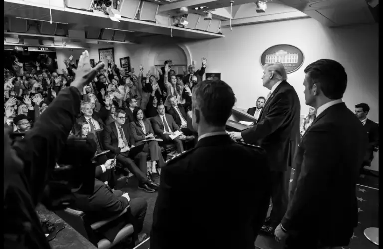 A black and white image of a formal press conference. President Trump at a podium faces journalists seated with raised hands, engaging with anticipation, addressing the Iran War and Ceasefire.