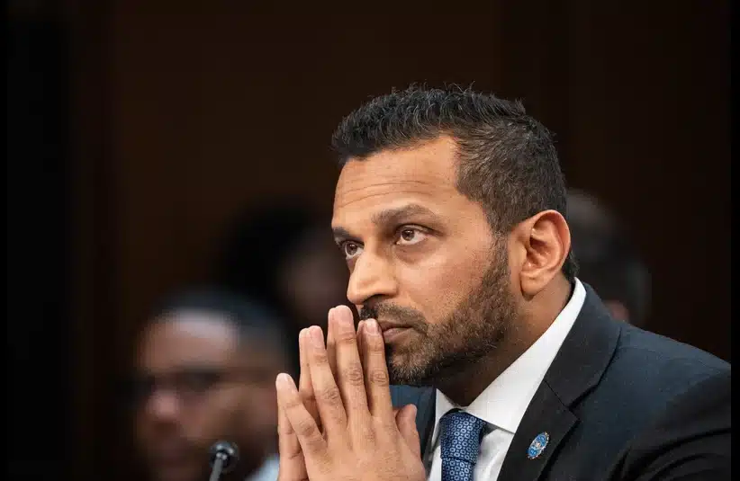Kash Patel, man in a suit sits with hands clasped, appearing focused and thoughtful. The background is blurred, suggesting a formal or professional setting.