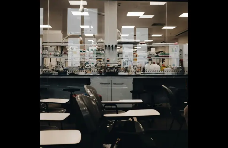 A brightly lit science lab viewed through glass, with shelves of equipment and supplies. Empty black chairs with white desks are in the foreground. America's science brain drain will have dire consequences.