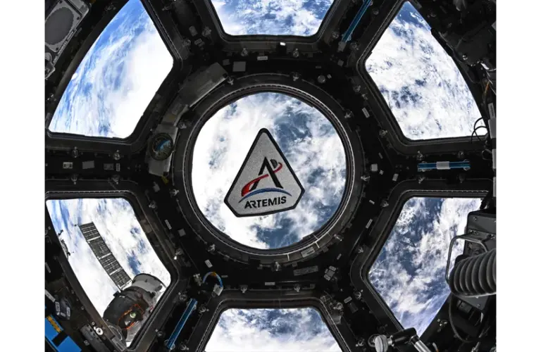 View of Earth from space through the circular window of a spacecraft. The Artemis II logo overlays the scene, framed by the structure, conveying a sense of exploration.