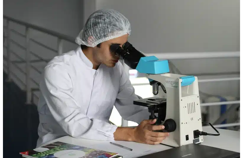 A scientist in a lab coat and hairnet examines samples through a microscope in a laboratory, suggesting focused scientific research and discovery. Important Human Clinical Trials Fentanyl Vaccine.