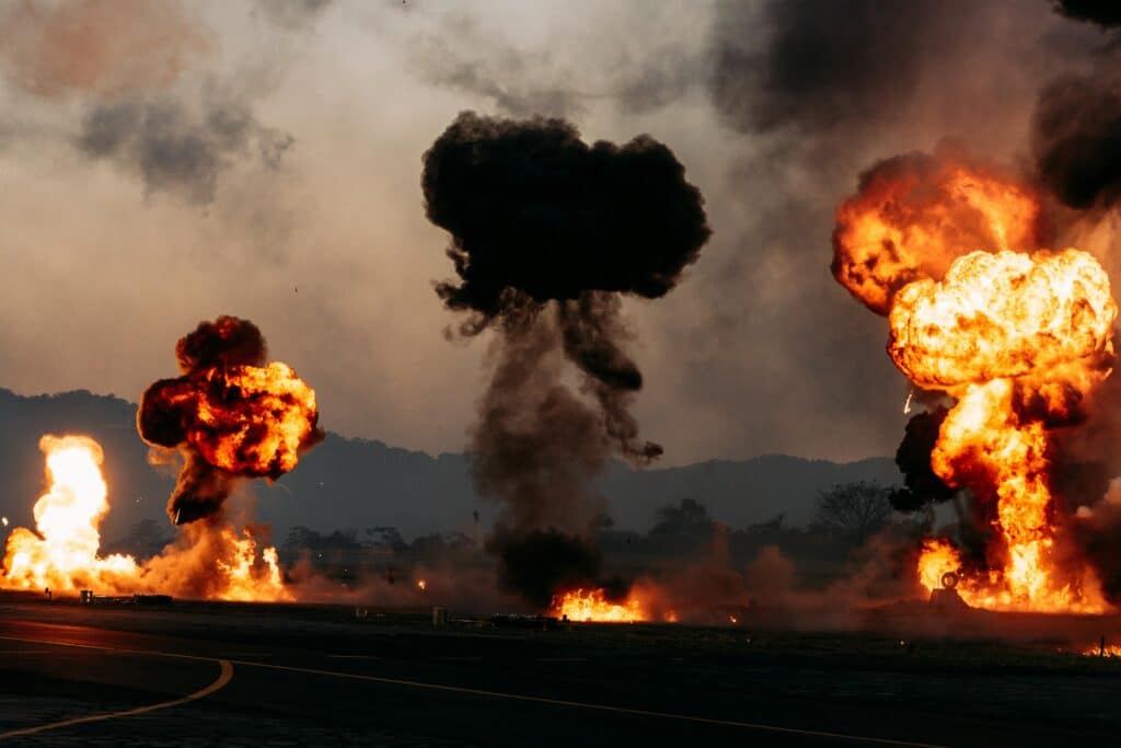 Explosive hostilities scene of multiple large explosions with billowing smoke and bright orange flames. The landscape is dark, conveying intense action.