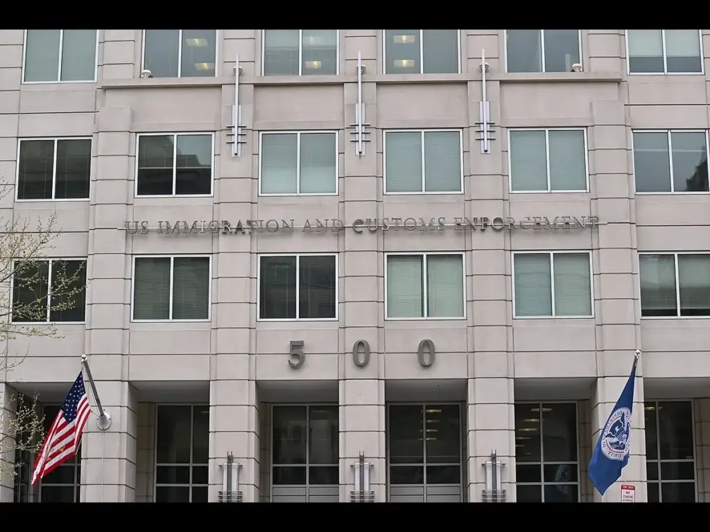Front view of a gray office building with "U.S. Immigration and Customs Enforcement" signage, two flags flanking the entrance, and large "500" numbers displayed. ICE building