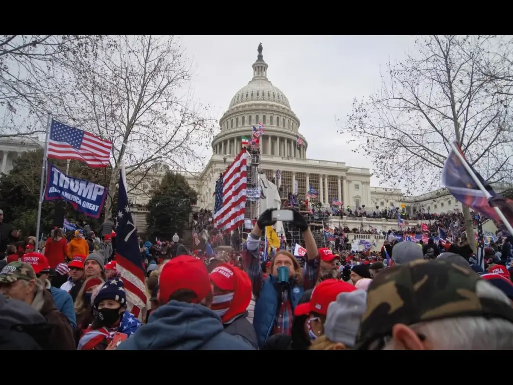 A large crowd gathers outside the U.S. Capitol, many wearing red hats and holding American flags and pro‑Trump flags, with people filling the steps and surrounding area during a political demonstration.