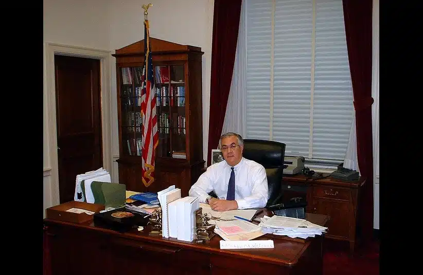 Barney Frank in white shirt and tie sits at a cluttered wooden desk with papers, books, and an American flag nearby. The office has white walls and red curtains. Trailblazer for politics.