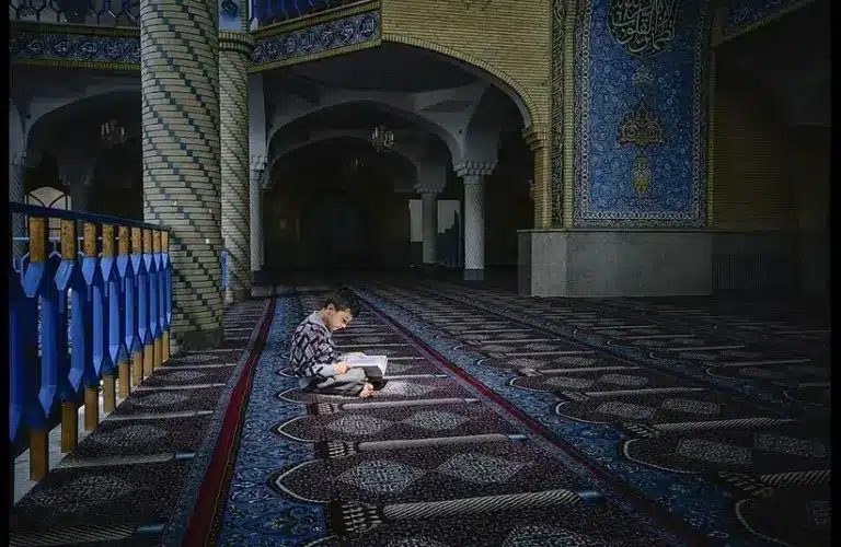 A young boy sits cross-legged reading a book on a richly patterned carpet inside a mosque. The space is serene, with blue and gold tiles adorning the walls. Recently there has been a shooting in San Diego leaving 3 dead, plus 2 more (the suspects)