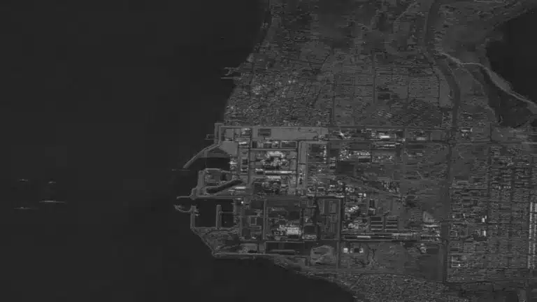 Aerial view of Bushehr Nuclear Power Plant in Iran, industrial coastal city at night. Dense grid of streets and buildings illuminated by artificial lights contrasts with dark sea. Urban landscape.