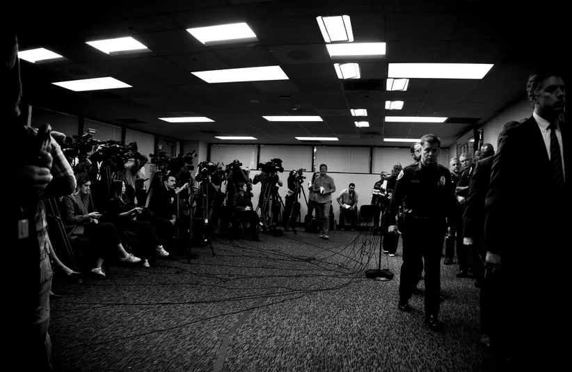 Black and white photo of a crowded press conference room filled with photographers and video cameras. A somber, professional atmosphere prevails.