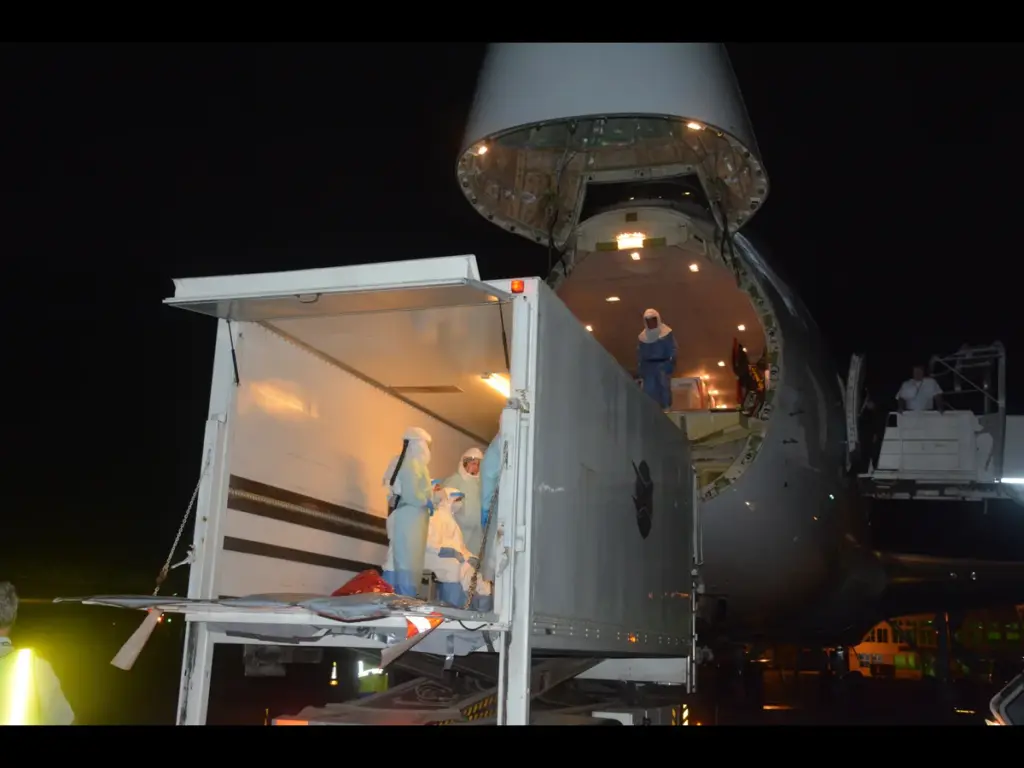 Cargo plane with open nose, loading at night. Workers in protective gear handle a container. Scene is lit, with a focused, meticulous atmosphere. Transportation of an Ebola patient.