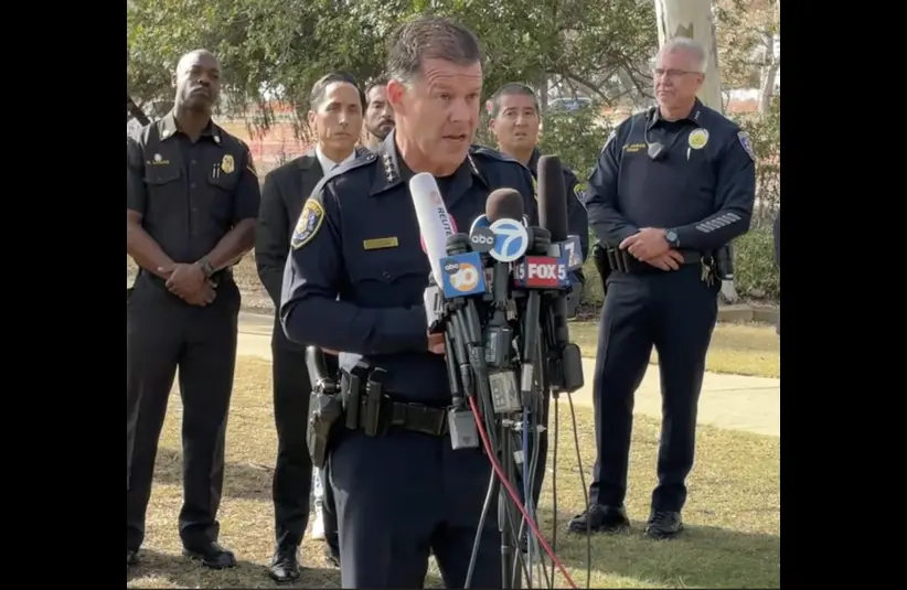 San Diego Police Chief Wahl, with fellow police officers in uniform, speaks at a press conference outdoors, surrounded by other officers. Microphones and news logos are in the foreground. This is an update on the San Diego Mosque Shooting.