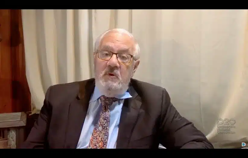 An older man, former Rep. Barney Frank, in a suit with a patterned tie and glasses speaks in front of a beige curtain. The setting is formal, conveying a serious tone.