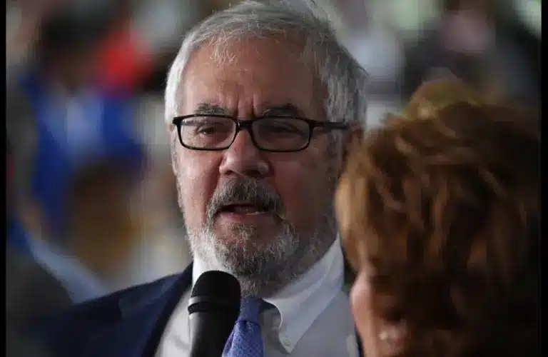An older man with glasses and a beard speaks into a microphone held by a woman, against a blurred, busy background. The tone is serious and engaged, former Rep. Barney Frank (D-Mass) speaking Democrats during interview