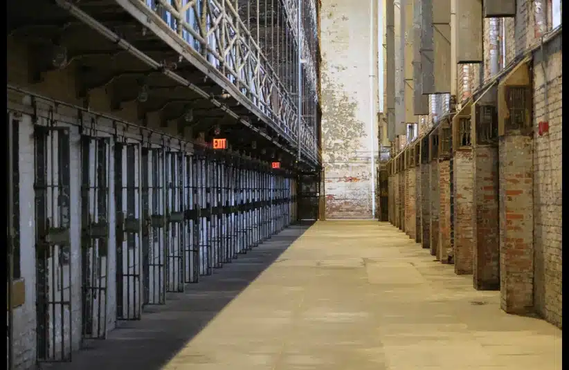 Dimly lit, long prison corridor with rusty barred cells lining the left; the concrete floor and exposed brick walls create a stark, somber atmosphere. Representative of a jail in New Orleans where ten inmates escaped. The sheriff is now being indicted.