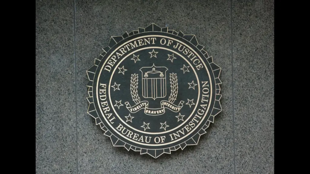 A circular FBI seal mounted on a textured stone wall, featuring a central shield with laurel branches, a red banner reading “Fidelity, Bravery, Integrity,” and outer text identifying the Department of Justice (DOJ) and the Federal Bureau of Investigation. They have removed Jan 6 files.