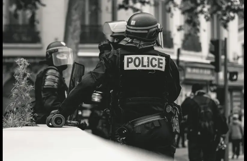 Black and white image of police officers in riot gear, standing on a street. One officer faces away, with "Police" on his back. Urban setting, tense mood.