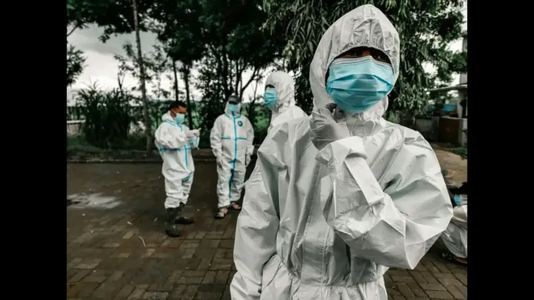 Five people wearing white protective suits and blue face masks stand on a paved path outdoors, surrounded by trees, conveying a serious tone. Items used for infectious disease protection, such as Ebola or Covid.