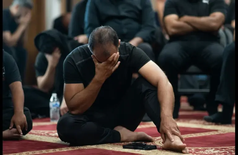 A man sits on a red carpet with a solemn expression, covering his face in his hand, surrounded by others in black clothing, conveying grief. His mood represents sorrow after the San Diego mosque shooting.