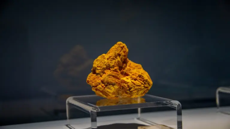 A vibrant uranium ore rock, irregularly shaped, displayed on a clear stand against a dark background. The setting is minimalistic and focused.