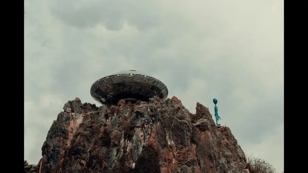 A UFO-shaped structure sits atop rugged rocks against a cloudy sky, with a small, green alien figure nearby, creating a mysterious, surreal atmosphere.